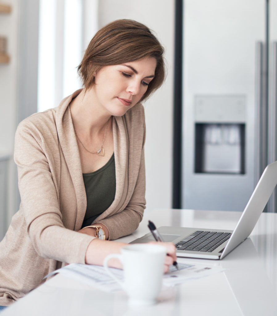 Illustrative image: women sitting at a table by her computer with a coffee mug. She is taking notes.